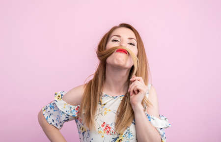 Funny brunette woman in dress holding hair on face like mustache while pointing up and looking at the camera over pink background.の写真素材