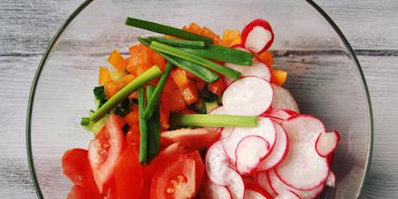 Glass bowl with cut vegetables for a vegan salad on the white wooden kitchen table. Radishes, tomatoes, bell pepper and cucumber. Close up. Wide photo.の写真素材
