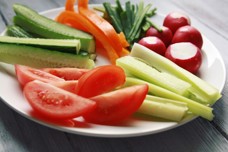 White plate with vegetables for a vegetarian salad. Radishes, tomatoes, celery, bell pepper, onion and cucumber. White wooden kitchen table. Close up.の写真素材