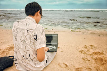Asian man trading on market online with his laptop in the front of the blue ocean