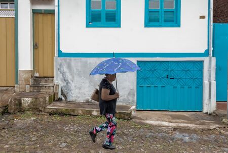MARIANA, MINAS GERAIS, BRAZIL. DECEMBER 23, 2019:  Unidentified woman walks with an umbrella along a cobbled streets with colouful houses in the background in Mariana, Minas Gerais, Brazil. Mariana is the oldest city in the state of Minas Gerais, Brazil. のeditorial素材