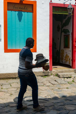 PARATY, RIO DE JANEIRO, BRAZIL - DECEMBER 28, 2019: A local man carries a pile of hats in front of colorful colonial buildings in the traditional city of Paraty, Brazil. The preserved colonial center of the city was included on UNESCO World Heritage list のeditorial素材