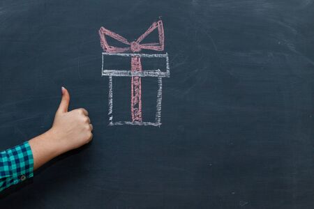 hand giving thumbs up on blackboard. Close up of thumbs up hand sign gesture of approval on chalkboard texture background with a picture of the box with the gift. Giftの写真素材