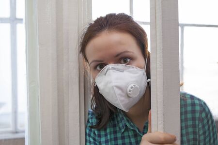 A girl in a medical mask looks out of an open door. Coronavirus epidemic, home quarantine during the COVID-19 infection.の写真素材