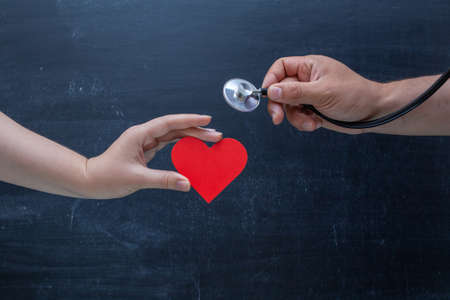 The girl holds a red heart symbol in her hand, the doctor's hand with a stethoscope