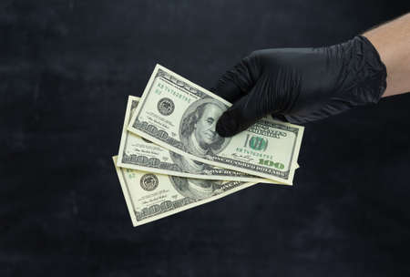 A man's hand in black medical gloves holds three hundred dollar bills in his hand