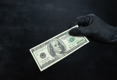 A hand in a black nitrile glove holds a hundred dollars against a dark background
