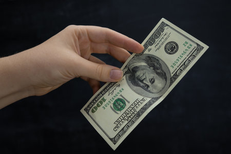 A woman's hand holds a hundred-dollar bill. Dark background
