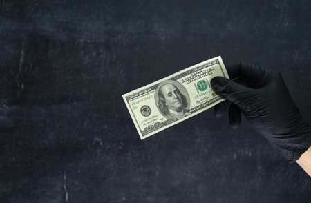 A hand in a black nitrile glove holds a hundred dollars against a dark backgroundの写真素材