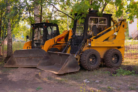 Two mini loaders with a bucket stand in the shade of a tree