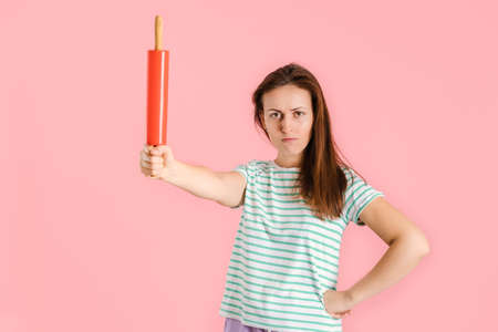 A beautiful brunette holds a red rolling pin at arm's length and angrily looks at the camera with...