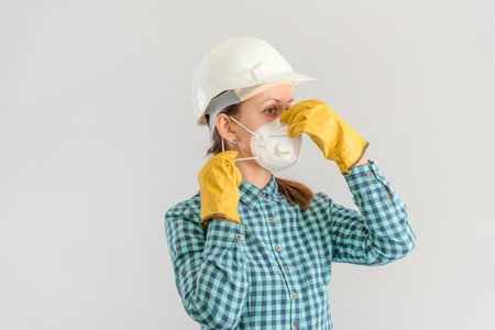 A young adult female construction worker puts on a protective medical mask. A female engineer in a hardhat wearing an N95 respirator and yellow gloves. Horizontal photoの写真素材