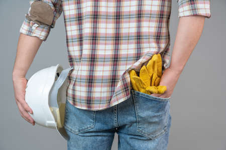 Close-up of a man from behind holding a protective helmet and work gloves in the back pocket of his pants on a gray background.の写真素材