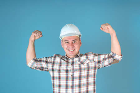 A smiling, sturdy man in a white helmet shows off his muscles. Builder shows biceps standing on a blue backgroundの写真素材
