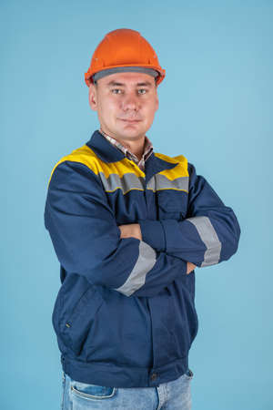 A worker in an orange helmet stands with his arms crossed on a blue background, vertical photoの写真素材