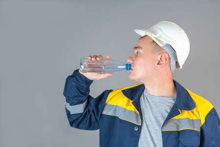 A construction worker in a white helmet and work clothes drinks water from a plastic bottle