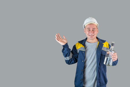 Drunk construction worker in work clothes and hardhat holds a bottle of alcohol in his hand and spreads his hands, copyspaceの写真素材