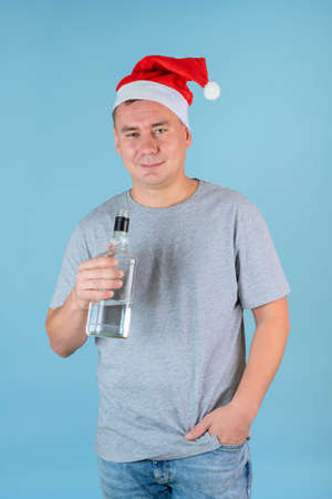 A drunk man in a New Year's Santa hat holds a bottle of alcohol standing on a blue backgroundの写真素材