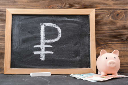 The symbol of the Russian currency is the ruble on a black chalk board in a frame, next to a piggy bank and paper billsの写真素材