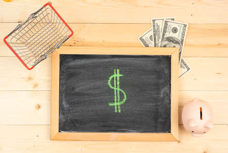 The Dollar symbol. A drawing of the dollar sign, on a black chalk board in a frame, next to dollar bills, a shopping basket and a piggy bank. Top viewの写真素材