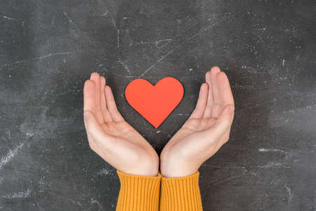 Female palms embrace a red heart-shaped figure, top view. Selective focus