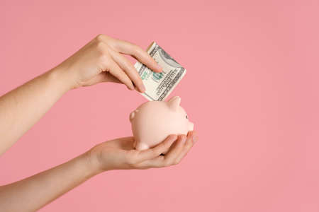 women's hands put a hundred-dollar bill folded in half in a piggy bank, pink background copyspaceの写真素材
