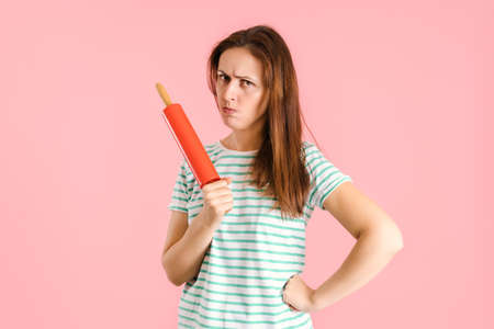 The angry brunette pursed her lips in displeasure and threateningly holds a rolling pin in her hands with her hand on her side. Studio shot on a pink backgroundの写真素材