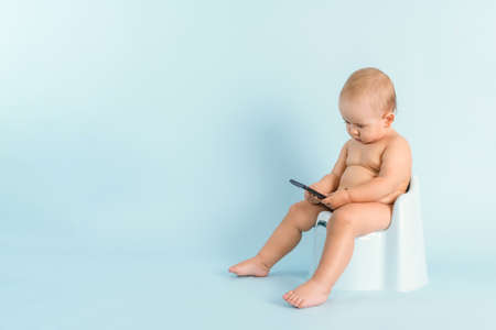 A newborn baby with a phone is sitting on a potty on a blue background. A child of 12-15 months is watching videos on a smartphone while sitting on a pottyの写真素材