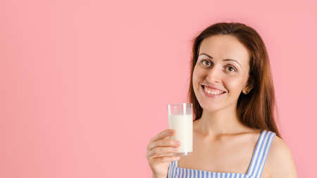 A smiling girl in a blue dress holds a glass of milk in her hands standing on a pink background. Close upの写真素材