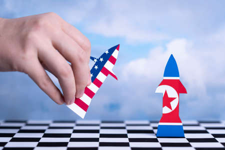 Hand holds a rocket with the US flag pointing towards a rocket figurine with the flag of North Korea. Chess boardの写真素材
