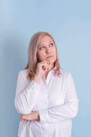 40s woman looks up thoughtfully holding her hand to her chin while standing on a blue background
