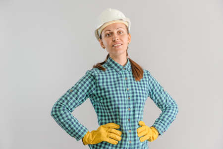 A young adult female construction worker in a white construction helmet stands with her hands in yellow gloves on her hips on a gray backgroundの写真素材