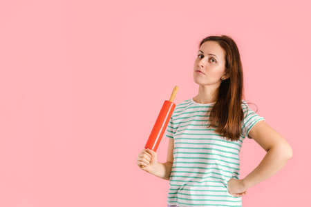 A beautiful brunette holds a red rolling pin in her hand and angrily looks at the camera. Studio shot on a pink backgroundの写真素材