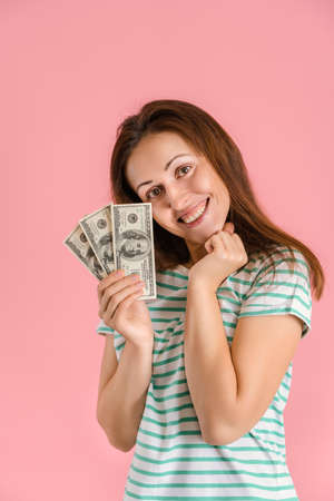 A joyful middle-aged girl smiles holding a fan of hundred-dollar bills in her hands standing on a pink backgroundの写真素材