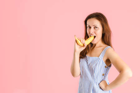 A brunette woman bites a yellow banana with a peel standing on a pink backgroundの写真素材