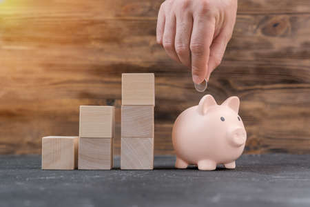 A man's hand puts a coin in a piggy bank, next to a stack of wooden cubes in the form of a ladderの写真素材