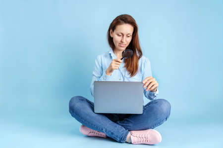 Search for information on the Internet. A young adult girl is sitting with a laptop on her lap and holding a magnifying glass in her hands on a blue backgroundの写真素材