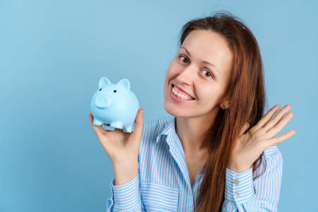 A happy young adult girl smiles holding a blue piggy bank in her hands on a blue backgroundの写真素材
