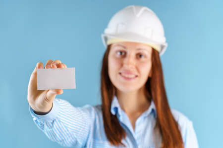 A female engineer in a hardcat holds a white business card at arm's length. Blue background, selective focusの写真素材