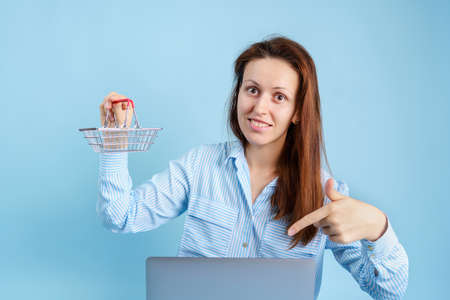 Online shopping on the Internet. A young adult girl with a laptop holds a shopping basket in her hands on a blue backgroundの写真素材