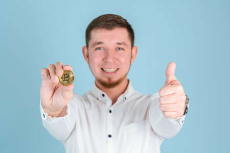 A smiling businessman holds a bitcoin gold coin at arm's length and shows a thumbs up gesture selective focusの写真素材