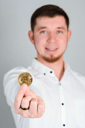 A bearded 30-year-old man in a white shirt holds a Bitcoin gold coin at arm's length, selective focusの写真素材