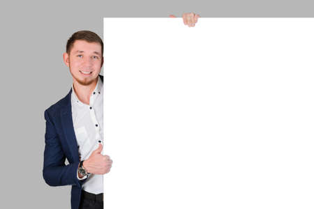 A smiling bearded man shows a thumbs up gesture while holding onto a white blank board, standing on a gray backgroundの写真素材