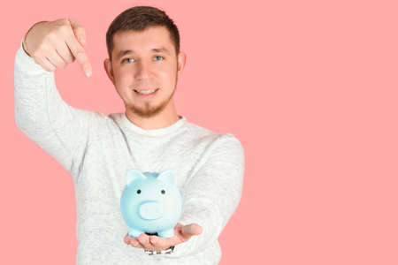 A red-haired, bearded man in a white sweater holds a blue piggy bank at arm's length and points at it with his finger. pink background with copyspaceの写真素材