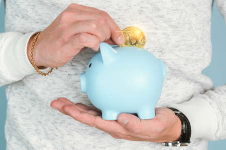 A man puts a bitcoin gold coin in a blue piggy bank, close-up