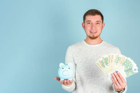 A red-haired bearded man holds a blue piggy bank and a fan of euro paper money, copyspace blue backgroundの写真素材