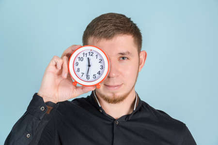 A young adult bearded man holds an alarm clock near his face standing on a blue backgroundの写真素材