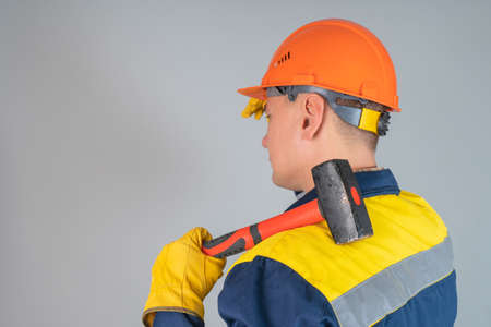 A worker in an orange helmet and with a hammer on his shoulder stands with his back to the camera on a gray background.の写真素材