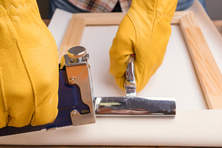 The artisan's hands in yellow protective gloves pull the canvas onto a wooden stretcher using a stapler pistol and forceps.の写真素材