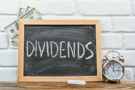The word DIVIDENDS on a black chalkboard in a frame standing on a white brick wall. Dollar bills and an alarm clock nearbyの写真素材
